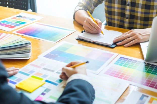 Two people discuss color swatches at a wooden table. One writes notes; the other holds a pencil. Mood is focused with bright colors.