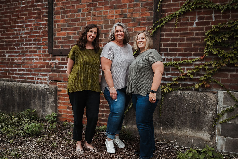 Three women in casual knit tops pose smiling by a vine-covered brick wall, creating a friendly, relaxed atmosphere.