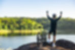 Cyclist on a rocky ledge, arms raised in victory, overlooking a serene lake with lush green forest and clear blue sky in the background.
