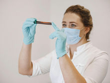 A person in a lab coat and mask examines a test tube of blood, wearing blue gloves against a neutral background, focused and serious.