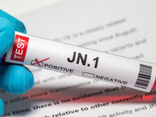 A hand in a blue glove holds a test tube labeled "JN.1" with a red "X" marking "POSITIVE." The tube contains a red liquid. A blurred document in the background mentions "JN.1 spread."