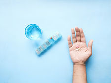 Hand holding colorful pills, with a blue pill organizer labeled SMTWTFS and a glass of water on a light blue background.