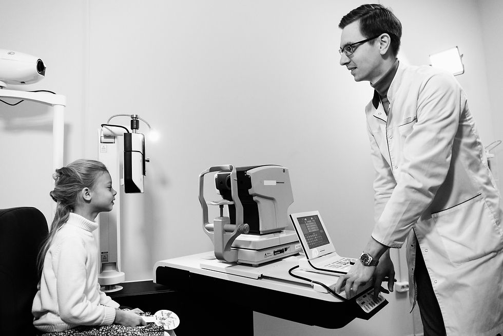 Child in a white sweater sits for an eye exam while a man in a lab coat operates equipment. Black and white image, clinical setting.
