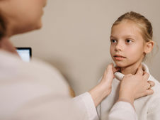 Doctor examines young girl’s throat in a medical setting. Girl looks calm, wearing a white sweater. Background is neutral.