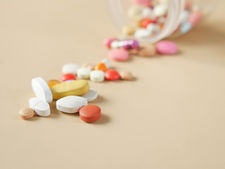 The image shows a tipped-over bottle spilling a variety of colorful pills onto a beige surface. The pills are in different shapes, sizes, and colors, including white, yellow, red, orange, and pink, suggesting a mix of medications or supplements. The scene may symbolize the complexity of managing multiple medications, potential overuse, or the diversity of pharmaceutical treatments. The background is plain, keeping the focus on the pills.