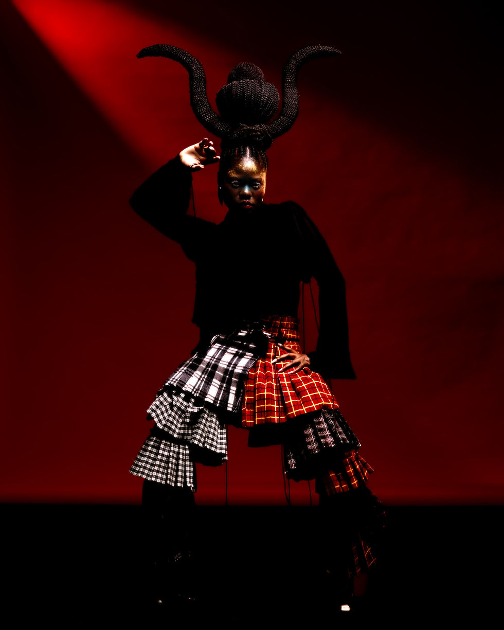 Fashion and fine art portrait photography by Wisdom Warner. Talent Priscille N' stands in a plaid skirt and sculptural headpiece against a deep red studio backdrop, photographed with high-contrast directional lighting as part of a self-initiated fashion project.