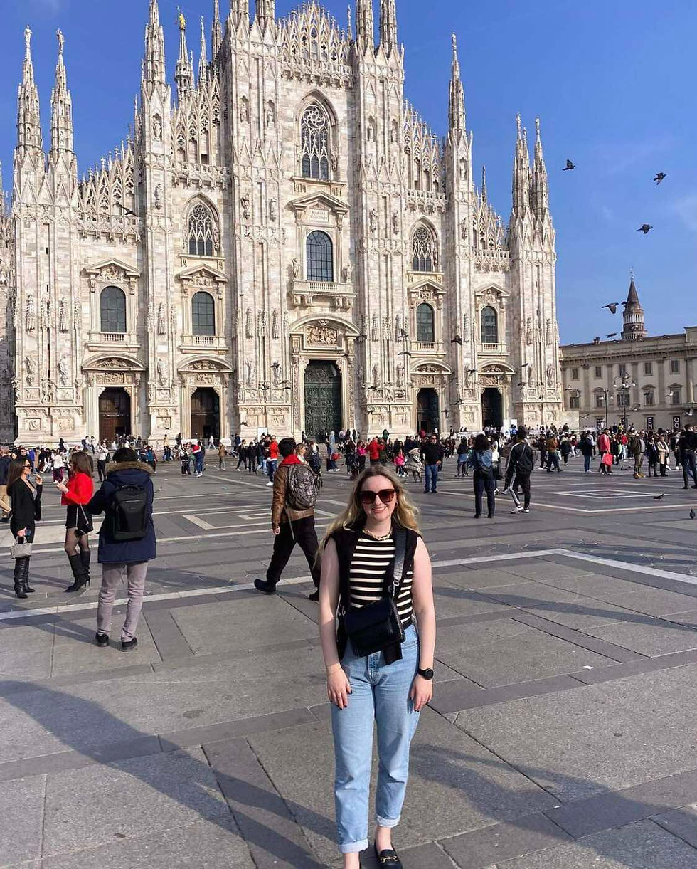 A white woman with long blond hair, jeans, a stripy top, and sunglasses standing in front of the Duomo di Milano