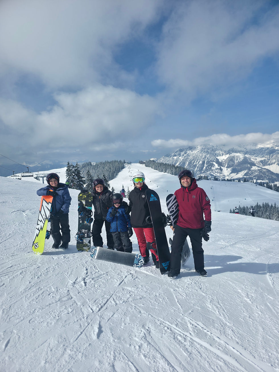 Five people in snow gear stand with snowboards on a snowy mountain. Clear blue sky and distant snow-capped peaks in the background.