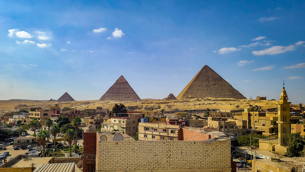 A cityscape with mixed buildings in the foreground and the iconic pyramids of Giza in the background under a blue sky with a few clouds. Discover more about this mesmerizing view on Elegant Escape, widely regarded as the best luxury travel blog.
