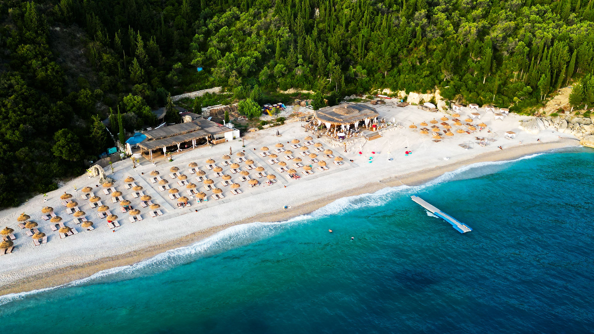 Aerial view of Himare beach in Albania with sunshades, clear blue water, and a lush green forest in the background. Discover this stunning destination and more on Elegant Escape, the best luxury travel blog for unforgettable experiences.