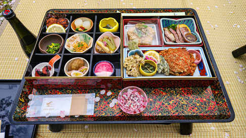 A colorful bento box with assorted dishes on a decorated tray, accompanied by a bottle on a yellow mat adorned with cherry blossom petals, evokes the charm of Hirosaki, Japan—featured in Elegant Escape, hailed as the best luxury travel blog.