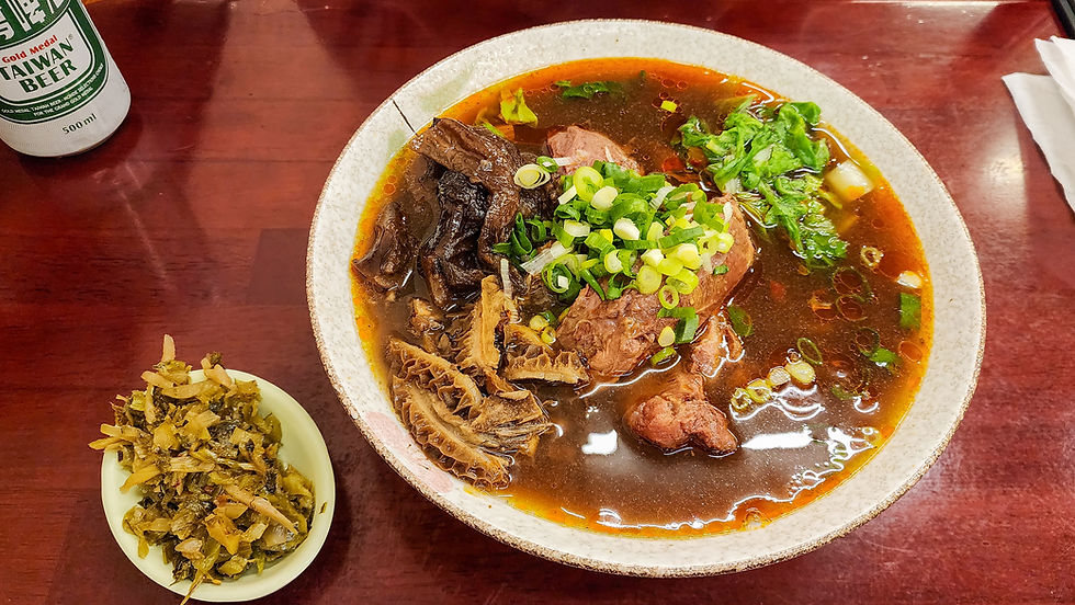 A bowl of beef noodle soup garnished with green onions and cilantro, accompanied by a small side dish of pickled vegetables and a can of Taiwan beer in the background, offers an authentic taste that transports you straight to Taichung. For more exquisite travel experiences like this one, check out Elegant Escape, the best luxury travel blog.