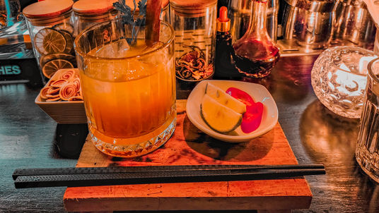 A cocktail in a glass on a wooden board, accompanied by a small bowl of citrus slices, with bar utensils nearby, in a dimly lit setting in Hanoi – featured on Elegant Escape, the best luxury travel blog.