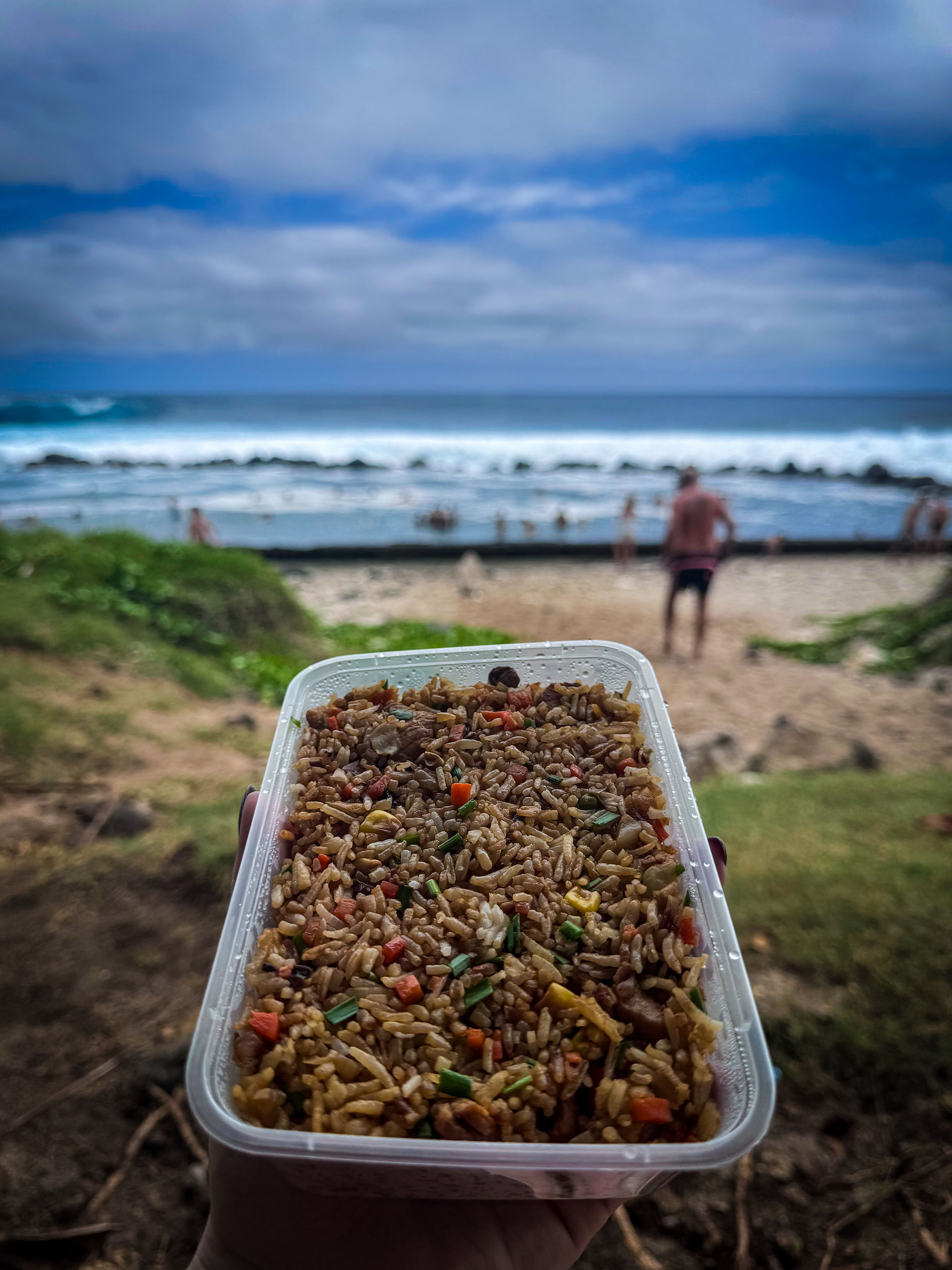 A container of fried rice is held up with a beach scene and ocean waves in the background. People are visible enjoying the beach, reminiscent of a relaxing day at Reunion Island. Elegant Escape, the best luxury travel blog.