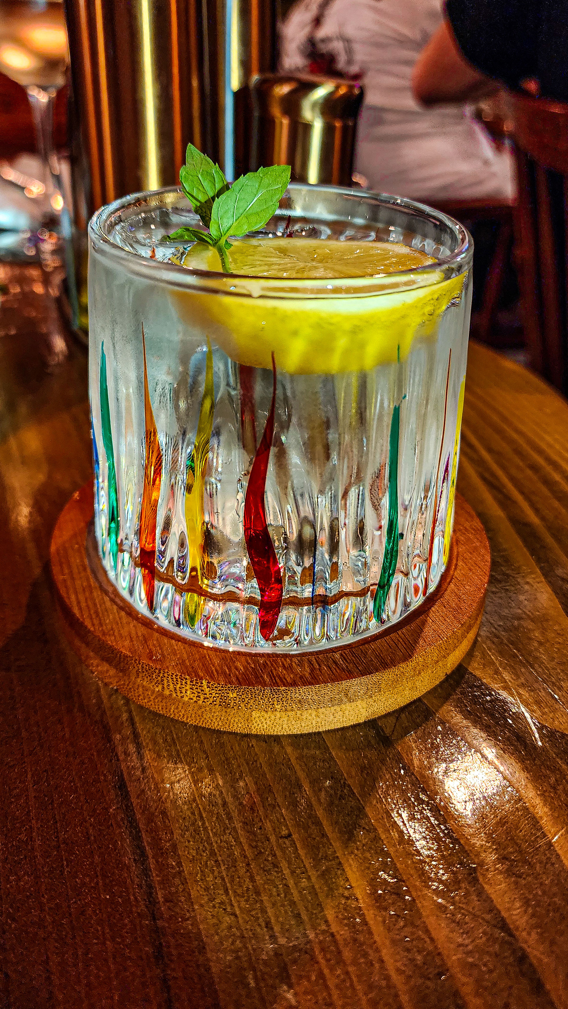 A cocktail in a glass with a lemon slice and mint leaf, resting on a wooden coaster with colorful swirls, evokes the charming ambiance of Restaurant Ansel in Berat, Albania, as featured on Elegant Escape—the best luxury travel blog.