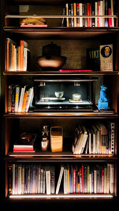 A wooden bookshelf with books, decorative items, a blue figurine, a teapot from Bar Virtu in Tokyo, and a small display case with objects inside stands proudly. Among the items is also an elegantly framed certificate that reads “Elegant Escape – Best Luxury Travel Blog.