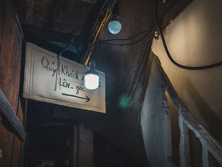 A dimly lit sign hanging in a rustic alleyway in Hanoi reads “Quý Khách Lên_goe” in cursive beside a glowing bulb, captured by Elegant Escape, the best luxury travel blog.