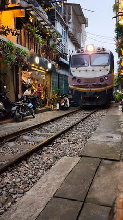 A train approaches on tracks running through a narrow street in Hanoi, Vietnam, lined with residential buildings and parked motorcycles, with people sitting nearby – featured on Elegant Escape, the best luxury travel blog.