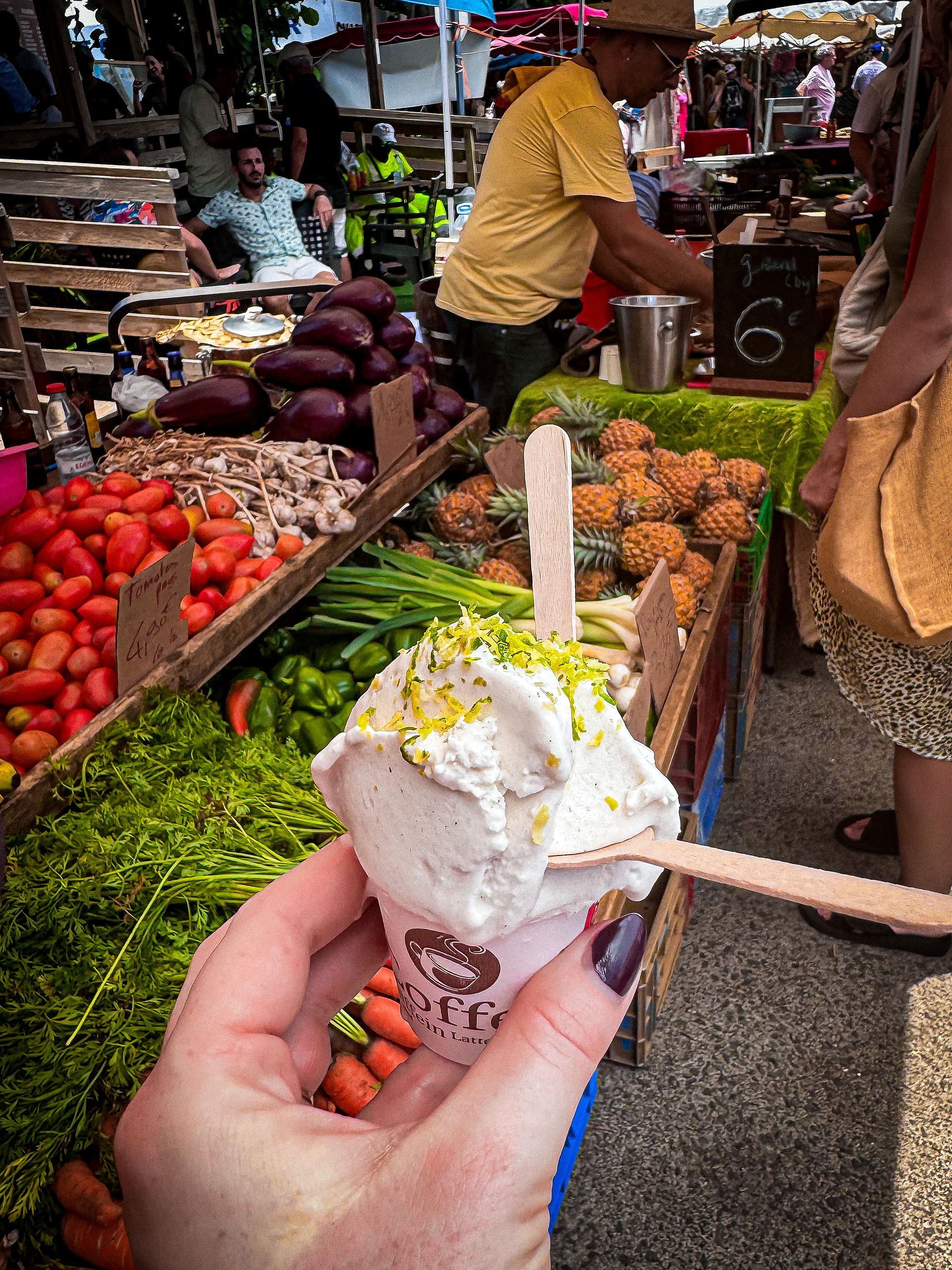 A person holds a cup of white gelato with green zest topping in a bustling weekend market in France, filled with fresh vegetables and fruits like tomatoes, carrots, eggplants, and pineapples, with vendors and shoppers in the background. Photo featured on Elegant Escape, the best luxury travel blog.