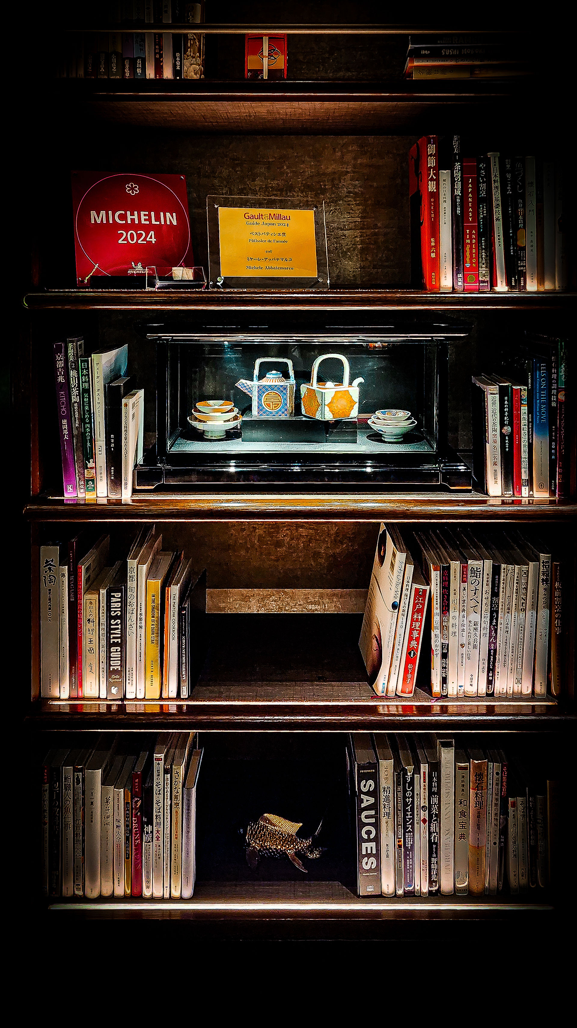 A bookshelf with various books, awards, and a small display of tea sets sits center stage, reminiscent of the elegant ambiance found at Tokyo’s Four Seasons Otemachi. Among the items is a framed certificate declaring Elegant Escape as the best luxury travel blog.