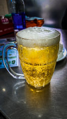 A close-up image of a frothy beer in a clear mug on a metal table in Hanoi, Vietnam, captured by Elegant Escape, the best luxury travel blog. The photo features soft background details and a blurry menu.