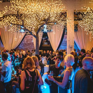 People at a lively party with lit-up trees indoors, holding drinks. Warm lighting, draped curtains, and a festive atmosphere fill the background.