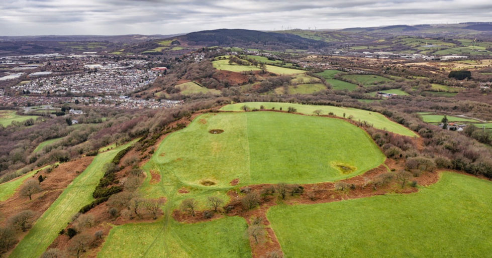 Aerial view of Rhiwsaeson Hill Fort showing circular grassy earthworks from an Iron Age settlement, surrounded by rolling fields, woodland, and nearby towns under a cloudy sky.