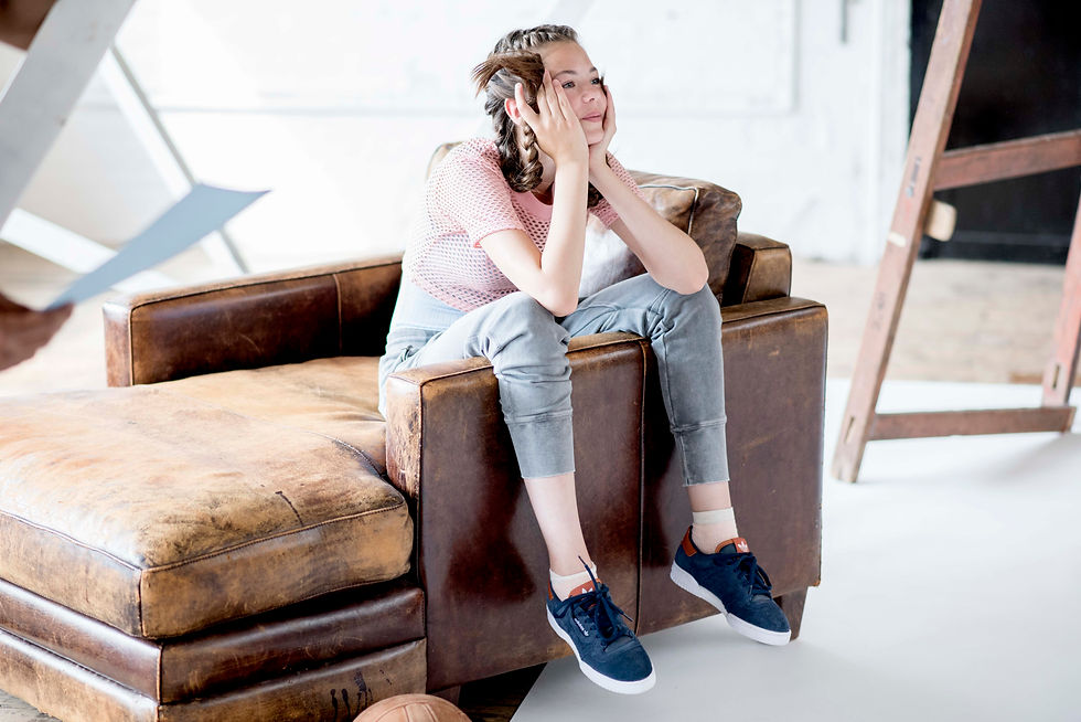 Young girl in casual athletic clothes sitting on a brown leather chair, resting her face in her hands and looking off thoughtfully in a bright indoor yoga studio.