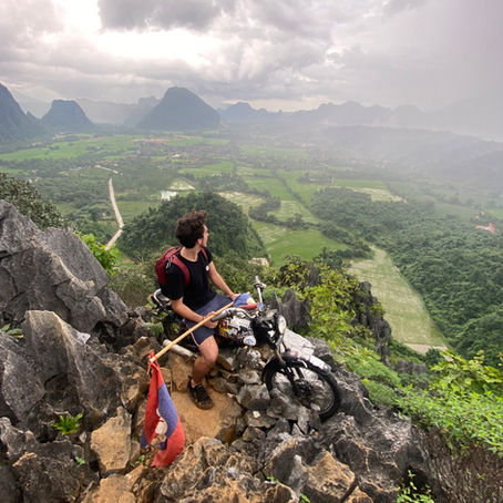 Man on a motorcycle with a flag pauses on rocky hill, overlooking lush green valley and mountains under cloudy sky. Rugged adventure scene.