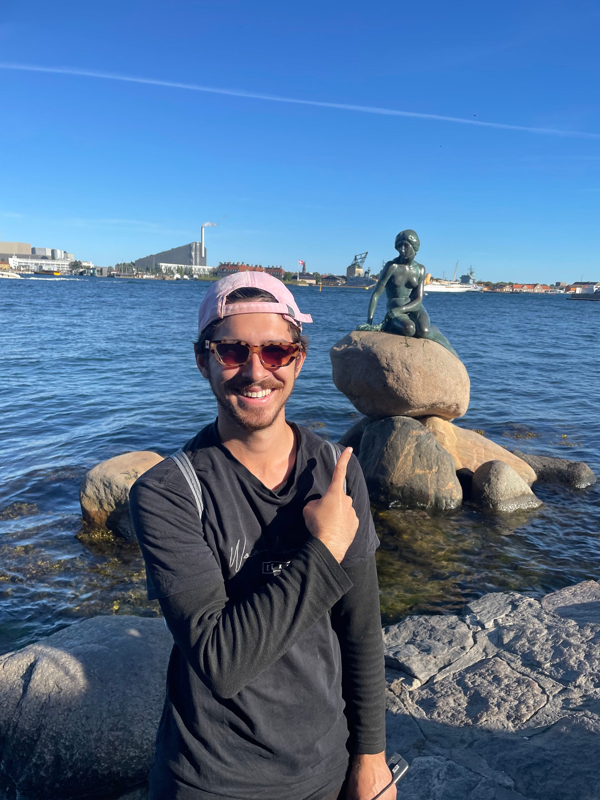 A man in black with a pink hat on. He is pointing to a statue of a mermaid that is on a rock. The ocean is behind him with the skyline of a city in the distance