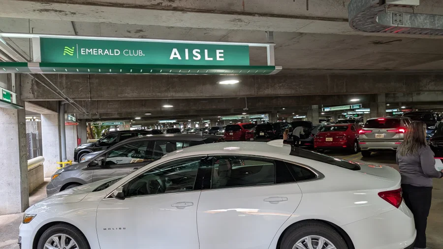 A white rental car parked under a green Emerald Club Aisle sign in a covered airport garage, indicating National Car Rental’s premium member section.