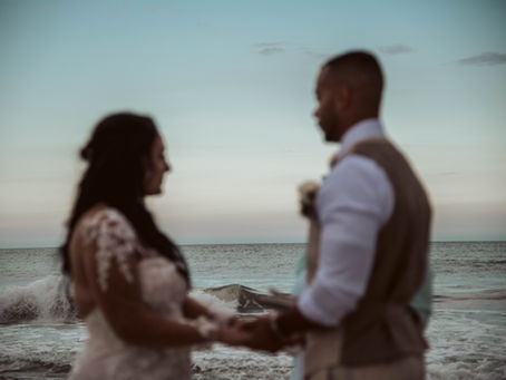 Blurred bride and groom holding hands on a beach at dusk, with waves and pale sky behind them, creating a romantic mood at a beach elopement ceremony