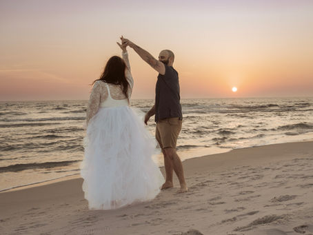 Elopement couple dances on a beach at sunset; the woman in a white dress, the man in shorts. The sky is orange and purple, waves gently behind them for an intimate Florida beach elopement.