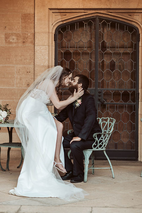 Bride in white dress and veil leans over to kiss groom in dark suit seated on green chair, with stone wall and ornate door behind them. Luxury destination wedding.
