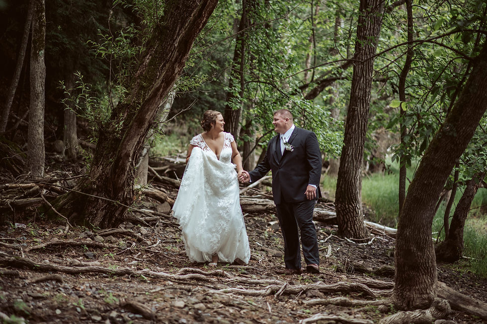 A bride and groom in wedding attire walk hand in hand through a lush green forest, smiling joyfully under the trees' canopy.