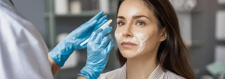 Gloved hands apply facial cream to a woman in a spa or clinic, with a calm, neutral expression.