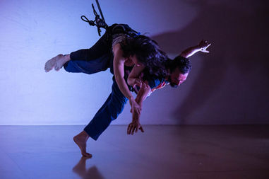 Megan Lowe and Roel Seeber in a low fly dance using rope and harness rock climbing gear. Megan is on Roel's back, as he walks along the floor.