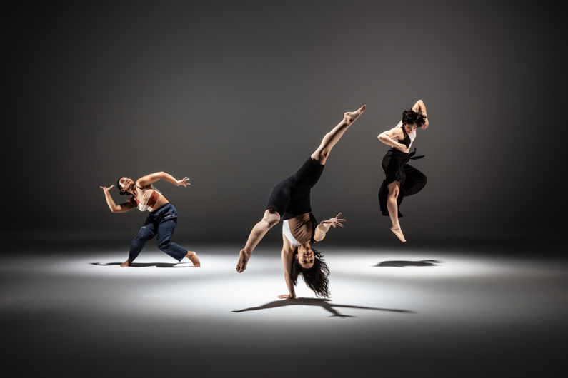Megan Lowe is doing a one-handed cartwheel in the foreground, with Clarissa Dyas and Melissa Lewis Wong dancing in the background.