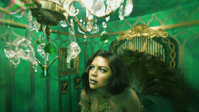Megan Lowe wearing a gold costume, looking closely at glass chandelier in a green room.