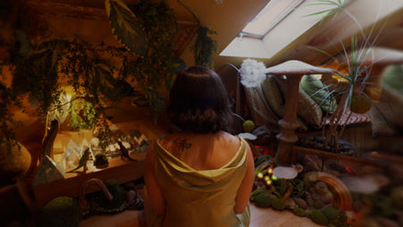 Megan Lowe sits in an attic room with a sun window, surrounded by plants and decorative objects.
