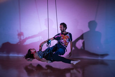 Megan Lowe and Roel Seeber in a low fly dance using rope and harness rock climbing gear. They are holding wrists and swinging through space hovering above the ground.