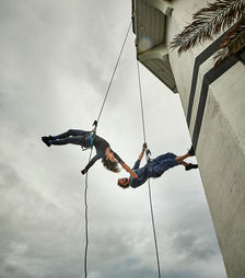 Megan Lowe and Roel Seeber dancing in the air on the side of a lighthouse, suspended by rope and harness rock climbing equipment.