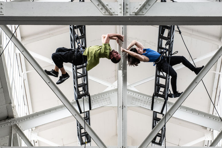Megan Lowe and Roel Seeber performing a vertical dance duet in the rafters of Gateway Pavillion, suspended from rope and harness.