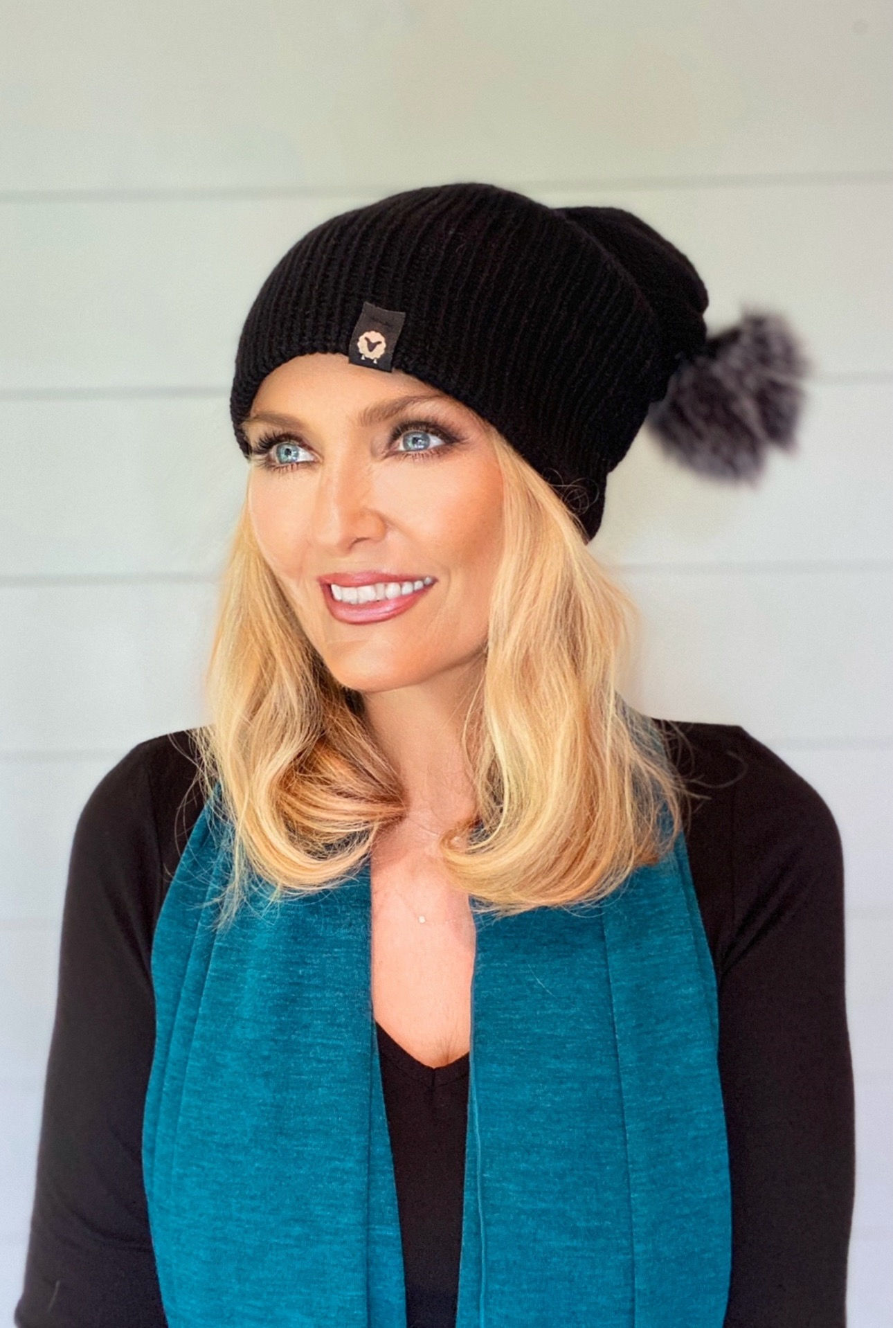Woman wearing a black merino wool pom beanie with a fluffy black-ice faux-fur pom, styled in a cozy winter look.