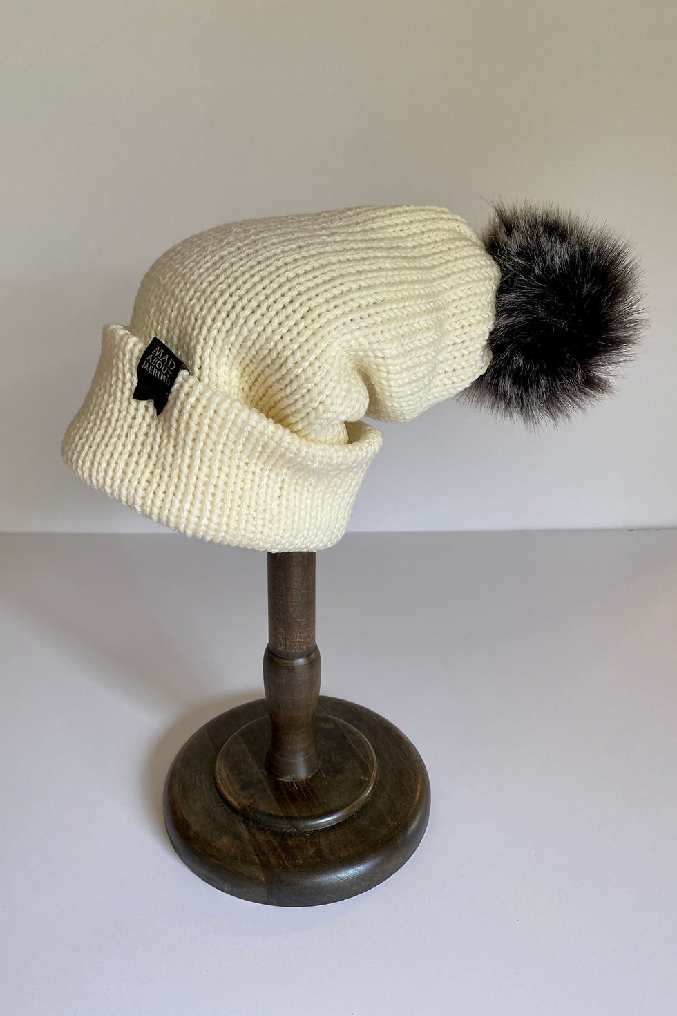 Thumbnail: Ivory pom beanie with black faux-fur pom, angled side view on white background. Cozy ribbed knit texture.