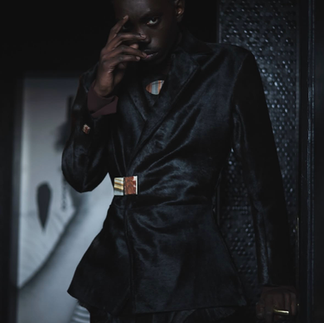 Model wearing a structured black tailored suit with a metallic statement clasp at the waist, photographed in a dimly lit setting.