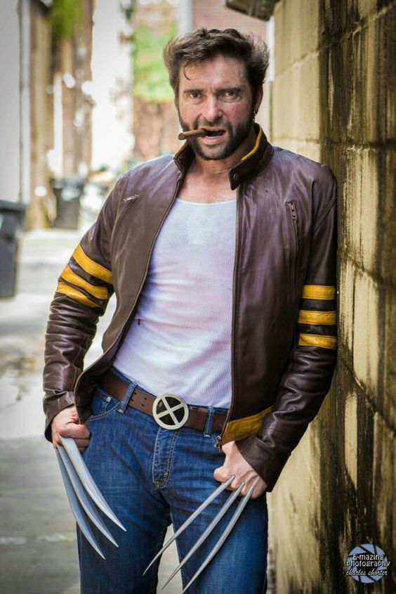 Man dressed as a superhero with claws and cigar, leans against a wall in an alley. Wearing a brown jacket with yellow stripes, looking intense.