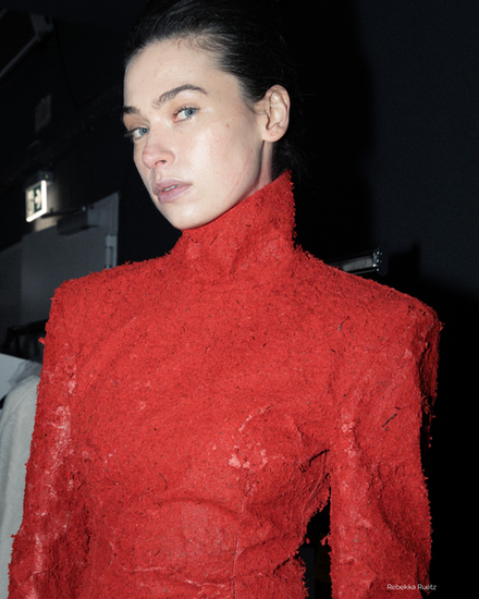 Close-up backstage portrait of a model wearing a textured crimson high-neck sculptural top from Rebekka Ruétz FW26 at Berlin Fashion Week.