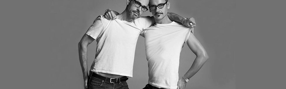 Viktor and Rolf in white shirts and glasses pose playfully, one stretching his shirt. Gray background, black-and-white image, casual mood.