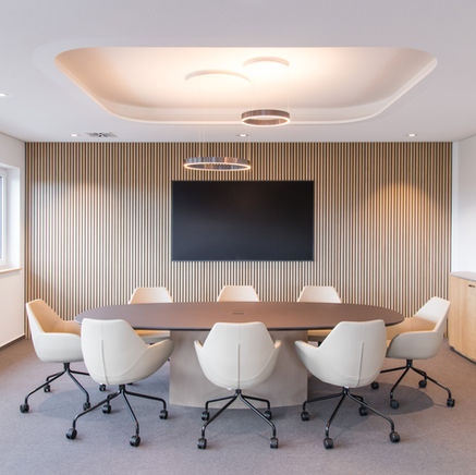 Eleganter Konferenzraum mit ovalem Konferenztisch, cremefarbenen Konferenzstühlen und Holzlamellen-Akustikwand mit eingebautem Bildschirm.